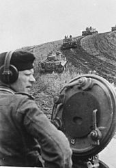 Photographie en noir et blanc montrant une colonne de chars montant une colline boueuse, l’avant-plan est occupé par un soldat qui émerge de la tourelle du char d’où est pris la photo.