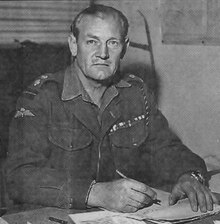 Black-and-white photograph of Churchill in uniform, sitting at a desk, and looking up at the camera while writing