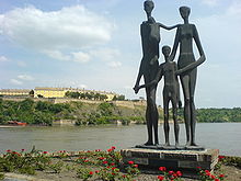 Au bord d'un fleuve, un monument composé de trois statues représentant des silhouettes émaciées.