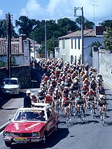 A road with, from front to back, a motor, a car, and tens of cyclists