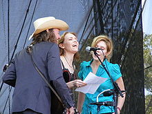 A man and two women crowded around a microphone and singing. The man is on the left and wearing a dark blue suit and a white cowboy hat. Welch, in the middle, is wearing a black dress, and the woman on the right is wearing a green dress.