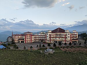 Photo of a cream-colored building with dark red highlights, with a larger section at the right capped by a brown pyramidal roof, and a narrower wing extending far off the to the left. The building is located on a hilltop with mountains in the background, and between the building and the camera lies a large, empty parking lot surrounded by a row of small decorative coniferous trees.