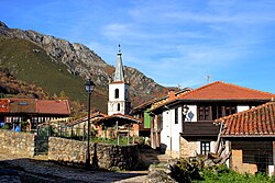 Image of the town, showing houses with red ceramic roof tiles, stone walls, a church, and the side of a mountain on the background.