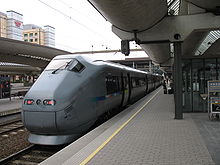 A sleek angular train at a modern train station, a yellow line is painted between the platform and the train