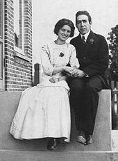 A young man in a suit and tie and a young woman in a light coloured dress sit on a stoop, holding hands