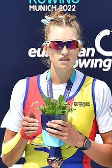 Amalia Bereș trägt den rumänischen rot-gelb-blauen Ruderanzug über einem weißen T-Shirt sowie eine Sonnenbrille. Um den Hals hat sie ein blaues Band, an dem die Bronzemedaille befestigt ist. Ihr rechter Arm ist bandagiert, in der linken Hand hält sie einen kleinen Blumentopf.