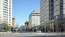 Ground-level view of the intersection of two wide streets, with one street one-way headed toward the viewer. The streets intersect within an urban area featuring high-rise office buildings on a sunny day during the winter.