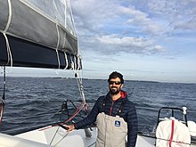 En buste, debout à la barre de son bateau. Barbe, lunettes de soleil, souriant.