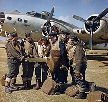 Photographie d'un groupe d'hommes en tenue de vol discutant devant des documents avec un bombardier en arrière plan.