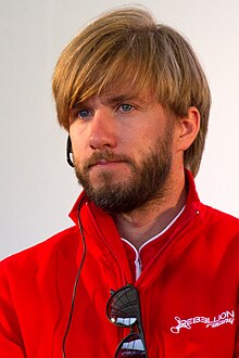 Photographie d'un pilote automobile, habillé en rouge, barbu, aux cheveux châtains, vu de face, en gros plan.