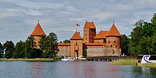 Frontale Farbfotografie einer braunen Burg mit vier Türmen. Die vorderen drei Türme sind mit zwei überdachten Gängen und kleinen Fenstern verbunden. Auf dem mittleren Turm ist eine Fahne. Im Vordergrund ist eine Wasserfläche mit Segelbooten und rechts eine Brücke.