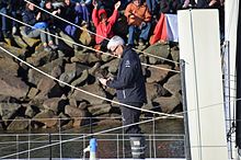 Cheveux blancs, lunettes, de profil. Debout sur son bateau, salué par la foule dans le chenal des Sables-d'Olonne, il prépare en souriant son téléphone portable pour filmer le public.