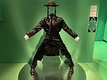 Costume de rabbin les bras et jambes écartés, exposé dans un décor vert.