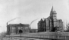 Two buildings stand side by side on a hill. The one on the left is two stories, with two smokestacks behind it. The one on the right is the larger, taller Tech Tower building.