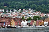 Foto einer am Ufer eines Gewässer gelegenen Siedlung. Direkt an der Küste befinden sich rote, orange, weiße und gelbe Holzhäuser. Dahinter steigt das Terrain an und es sind vor allem weiße Häuser zu sehen.