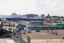 In this photograph, many large containers and other cargo are lined up in the city's ferry port. A ferry can be seen docked in the background.