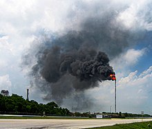 Vue au niveau du sol d'une haute torchère émettant un nuage de fumée noire, au milieu d'un ciel bleu. Au premier plan figure une route et des arbres sont présents à gauche de l'image.