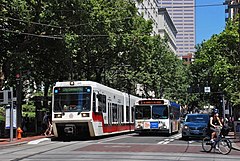 Photograph of a Max train on the left, a bus in the center and a woman on a bicycle making a turn off the Portland Transit Mall