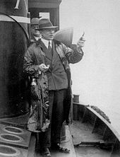 A man wearing hat and coat, gesturing with a pipe aboard a small ship