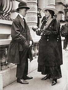 Mary Newcomb is standing on sidewalk selling tickets to a man with a hand in his pocket getting money out