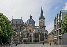 Seitliche Farbfotografie eines Doms mit mehreren Gebäudeteilen. Links steht das Kirchenschiff mit bodenlangen Kirchenfenstern, in der Mitte ein Kuppelbau mit verzierten Dachgiebeln und rechts ein Turm mit kleinen Ecktürmen. Im Vordergrund sind zwei kleinere Bauten mit langen Kirchenfenstern. Der Vorplatz wird an beiden Seiten von alten Häuserfassaden gesäumt und vorne rechts steht ein modernes Geschäftshaus.