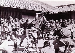 Photo d'un homme brandissant une épée, entouré d'autres hommes armés et menaçants.