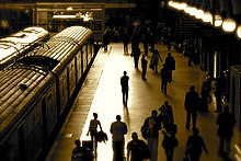 Un quai de gare vue en légère plongée, avec deux trains côte à côte à l'arrêt. La lumière du soir est vive et rasante et une silhouette en contre-jour se distingue au bord du quai, parmi la foule des passagers