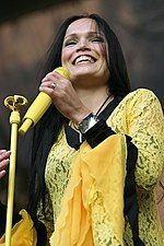 Photo en couleurs d'une femme souriante aux longs cheveux bruns, habillée d'un haut en dentelle jaune et noire et tenant dans sa main un micro jaune.