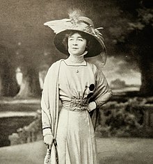 A white woman with dark hair, standing. She is wearing a bird-embellished hat with a wide brim, pearls, and a light-colored corseted gown with a high lace collar.