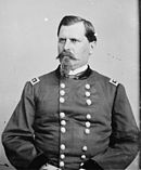 Black and white photo shows a stern-looking man with a moustach and pointy beard. He wears a dark military uniform with two rows of buttons.