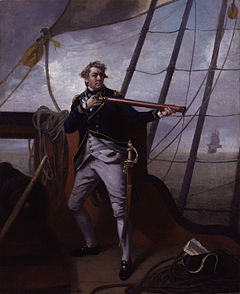 A large man in a blue uniform strikes a dramatic pose holding a telescope on the quarterdeck of a ship. In the distance another ship can be seen with its sails set.