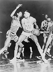 A basketball player, wearing a light-colored jersey with the number "13", is holding a basketball in front of another basketball player who is wearing a dark-colored jersey with the number "6".