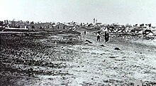 In the distance a group of people walk down a dirt road away from the village. The foreground shows vacant ground.