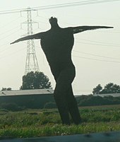 Large statue of a human figure with arms outstretched. In the background are trees and an electricity pylon.