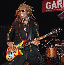 T.M. Stevens wearing black clothing, a variety of jewelry, and black sunglasses, standing onstage in front of a drum kit, playing bass guitar with mouth open