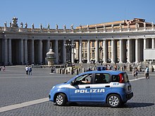 Farbfotografie von einem riesigen Platz, der von einem hellen Säulengang mit Figuren drauf umschlossen ist. Verteilt laufen Menschen auf dem Platz, das einen Springbrunnen hat. Im Vordergrund ist ein blau-weißer Polizeiwagen mit der Aufschrift „Polizia 113“ zu sehen. Im Wagen sitzt ein Mann, dessen Arm im Licht gut zu erkennen ist.