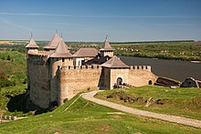 Farbfotografie mit Blick von einem Hügel auf die Festung mit einem Weg, der von unten rechts hinführt. Die gut erhaltene Festung mit seinen vier Türmen und einem Gebäude im Innenhof ist von hügeligen Wiesen umgeben. Auf der rechten Seite fließt der Fluss Dnister hinter der Festung vorbei. Man erkennt in der Ferne einen Ort.