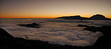 Coucher de soleil en fond avec un sommet. Au premier plan, une mer de nuages blancs.