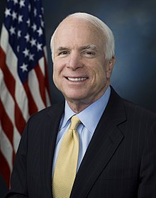 A portrait photograph of a smiling white-haired elderly man wearing a dark business suit, with American flag in the background.