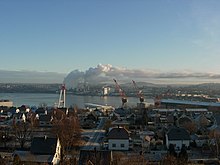 Foto einer Stadt, die von einem Gewässer in zwei Teile gespalten wird. Auf beiden Seiten sind Industrieanlagen wie Hafenkräne und Fabriken zu erkennen. Aus den Fabriken auf der gegenüberliegenden Uferseite entweicht Rauch.