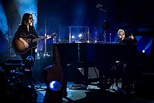 Gaga playing piano and Hilary Lindsey standing opposite to her paying guitar onstage.