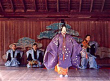 Acteur debout en tenue traditionnelle violette, masque blanc souriant. Trois musiciens visibles en arrière plan.