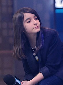 A white teenager with long brown hair is sitting, holding a microphone, wearing a dark blue shirt. She is leaning to her right, looking to her left—right of the camera.