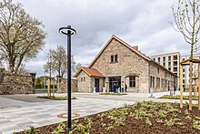 Eine Fotografie, auf der das neue Gelände des Quartiers Sankt Leonhard zu sehen ist. Im Vordergrund steht eine schwarze Laterne. Eine neu angelegte Bepflanzung mit einem grünen Bodendecker und einem jungen Baum mit langsam grüner werdenden Knospen ist im Vordergrund zu sehen. Im Mittelgrund liegt ein mit hell- und dunkelgrauen Platten gepflasterter Platz. Dahinter sind die restaurierten Scheunengebäude zu sehen, die moderne, schwarze Sprossenfenster haben. Eine ebenfalls aus schwarzem Stahl und Glas bestehende Eingangstür ist dem Betrachter zugewandt. Davor stehen vier Menschen an einem Stehtsich mit Husse. Im Hintergrund links sieht man einen Teil der Sankt Leonhard-Straße durch einen steinernen Torbogen hindurch, rechts erhebt sich dermoderne und neu gebaute sechststöckige Flachbau.