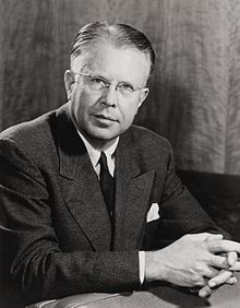 Head and shoulders of a man wearing rimless glasses, and a dark suit and tie