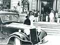 Entouré de personnages, Mussolini en uniforme blanc, au pied de l'escalier d'un hôtel de luxe, semble s'apprêter à monter dans une automobile sombre.