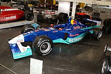 Photographie d'une monoplace de Formule 1 bleu foncé et vert clair, vue de trios-quarts, dans un musée.