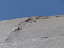 An inclined wall covered with white pieces of ceramic under a blue sky. Some of the tiles are missing, others are detached from the wall and raise against other pieces.