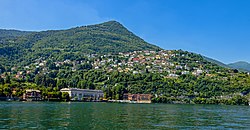 A mountain rising above water on a day with clear blue sky. There are large houses at the water's edge, and smaller houses higher up.