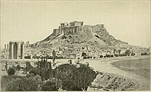 Old photograph of the Acropolis, with its ruined monuments, rising precipitously above a sparsely-built city.